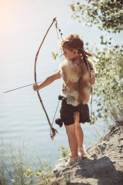 Kürk giysili ve saç bandı takmış genç bir çocuk, sakin bir orman ortamında ok ve yay ile gölün kenarında duruyor. Macera, tarih öncesi ve kültürel temalar için mükemmel.. 