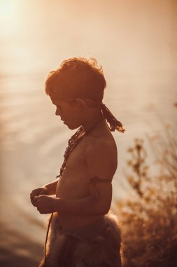 İlkel giysiler içinde genç bir kabile çocuğu, gün batımında ılık bir gölün kenarında duruyor, dingin ve zamansız bir siluet yaratıyor. Doğa, macera ve kültürel temalar için mükemmel. 