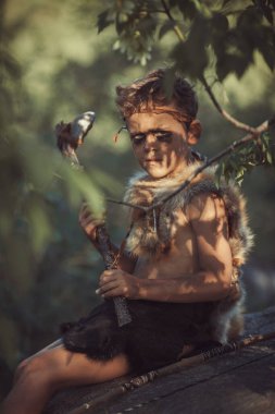 Yüzü boyalı ve kürk giysili genç bir çocuk ilkel bir baltayla ormanda oturuyor, doğal ışık ve yapraklarla çevrili. Tarih öncesi, macera ve kültürel temalar için mükemmel.. 