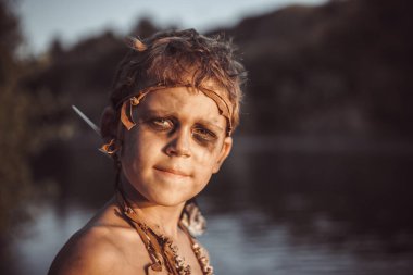 Kabile yüz boyası ve deri başlıklı genç bir çocuk gün batımında sakin bir gölün yanında duruyor. İlkel ve maceracı bir ruh yayıyor. Tarih öncesi ve kültürel temalar için mükemmel. 