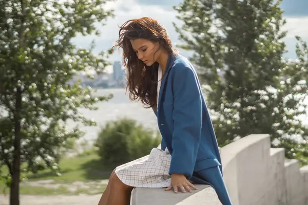 Uzun kahverengi saçlı ve mavi ceketli zarif genç bir kadın kayadan bir çıkıntının üzerinde düşünceli bir şekilde oturuyor. Kavramsal moda çekimi editoryal, promosyon veya yaşam tarzı reklam kullanımı için idealdir.