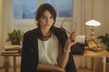 Beyaz bluz ve siyah ceket giyen, şık bir ofis odasında gözlük takan, sıcak ışıklandırmalı beyaz bir kadının yakın çekimi. İş, yaşam tarzı veya editoryal kullanım için ideal. 