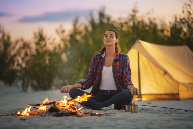 Huzurlu bir kadın kamp gezisi sırasında parlayan bir kamp ateşinin yanında meditasyon yapıyor. Arka planda çadır olan sakin bir açık hava ortamı, farkındalık, seyahat ve sağlık teması için ideal.. 