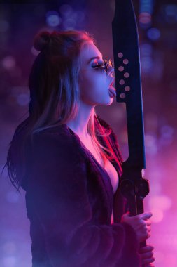 Baştan çıkarıcı siber punk kostümlü kadın güneş gözlüğü takıyor, neon ışıklı şehir manzarasına karşı kılıçla poz veriyor. Anime temalı promosyonlar için ideal, fütürist moda, ve sinir bozucu, modern reklamlar.