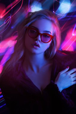 Güneş gözlüklü baştan çıkarıcı sarışın kadın, canlı neon ışıklarla poz veriyor ve dinamik bir zemin oluşturuyor. Moda tanıtımları, gece hayatı reklamları ve modern, uçuk kavramları için mükemmel..