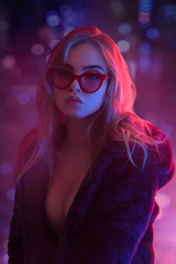 Baştan çıkarıcı, güneş gözlüklü, parlak neon ışığıyla yıkanmış, yakası açık sarışın bir kadın. Moda promosyonları, gece hayatı reklamları ve modern, keskin kavramlar için idealdir..