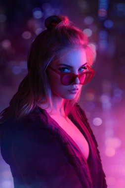 Baştan çıkarıcı, güneş gözlüğü takan, parlak neon ışığıyla aydınlatılmış sarışın kadın. Moda promosyonları, gece hayatı reklamları ve modern, keskin kavramlar için idealdir..