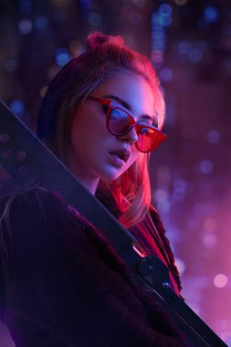 Kırmızı güneş gözlüğü takan ve elinde neon ışıklarla aydınlatılan bir kılıç tutan gelecekçi siber punk sarışın kadın. Bilim kurgu modası, gece hayatı promosyonları ve heyecanlı, kavramsal reklamlar için ideal..