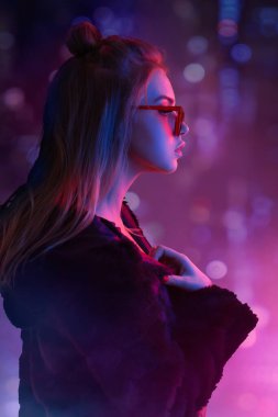 Şık, sarışın, güneş gözlüğü takan, parlak neon şehir ışıklarıyla aydınlatılmış bir kadın. Moda tanıtımları, gece hayatı reklamları ve modern şehir kavramları için ideal..