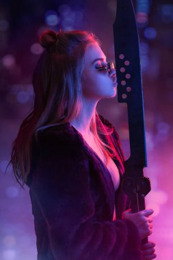 Baştan çıkarıcı siber punk kostümlü kadın güneş gözlüğü takıyor, neon ışıklı şehir manzarasına karşı kılıçla poz veriyor. Anime temalı promosyonlar için ideal, fütürist moda, ve sinir bozucu, modern reklamlar.