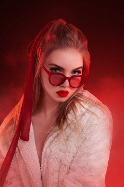 Kırmızı güneş gözlüğü takan şık sarışın kadın ve dramatik kırmızı ışıklandırmalı beyaz kürk manto. Moda promosyonları, tatil reklamları ve modern, uçuk kavramları için ideal..
