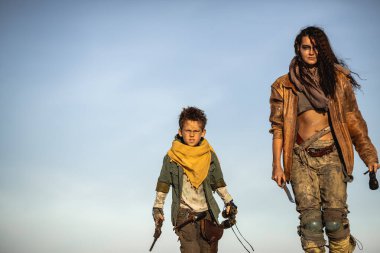 Kıyamet sonrası kostümlü silahlı kadın ve çocuk gün batımında çölde yürürler. Yüksek kaliteli bilim kurgu hayatta kalma konsepti fantezi tanıtımları, macera posterleri ve aksiyon öyküleri için mükemmeldir..