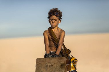 Kıyametten sonra çölün ortasında bir çocuk. Nükleer kıyamet sonrası patlaması. Kıyamet sonrası yaşam konsepti. Arka planda çöl ve ölü çöl var. Bıçaklı, gaz maskeli genç bir adam, göğsünü sürükleyen bir kupa.