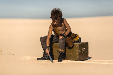 Kıyametten sonra çölün ortasında bir çocuk. Nükleer kıyamet sonrası patlaması. Kıyamet sonrası yaşam konsepti. Arka planda çöl ve ölü çöl var. Bıçaklı, gaz maskeli, kupa göğsünde oturan genç bir adam.