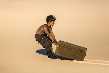 Kıyametten sonra çölün ortasında bir çocuk. Nükleer kıyamet sonrası patlaması. Kıyamet sonrası yaşam konsepti. Arka planda çöl ve ölü çöl var. Bıçaklı, gaz maskeli genç bir adam, göğsünü sürükleyen bir kupa.