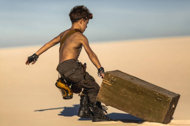 Kıyametten sonra çölün ortasında bir çocuk. Nükleer kıyamet sonrası patlaması. Kıyamet sonrası yaşam konsepti. Arka planda çöl ve ölü çöl var. Bıçaklı, gaz maskeli genç bir adam, göğsünü sürükleyen bir kupa.