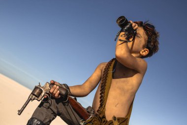 Kıyametten sonra çölün ortasında bir çocuk. Nükleer kıyamet sonrası patlaması. Kıyamet sonrası yaşam konsepti. Arka planda çöl ve ölü çöl var. Göğsünde silah taşıyan genç bir adam arıyor.