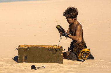 Üstsüz çocuk çölde diz çöküp yeşil askeri tarzda bir davadan alınan dürbünleri inceliyor. Yüksek kaliteli bilim kurgu hayatta kalma görüntüsü sinematik hikaye anlatma, fantezi tanıtımları ve vites temalı görseller için mükemmeldir.