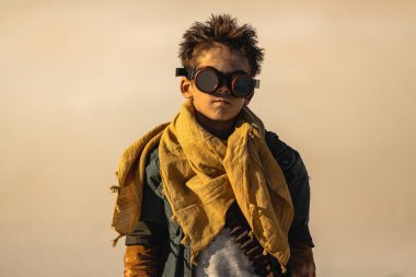 Engebeli hayatta kalma ekipmanları ve gözlükleri olan genç bir savaşçı çocuk sert güneş ışığı altında çorak bir çölde duruyor. Distopyan ve macera kavramları için mükemmel bir görüntü.. 