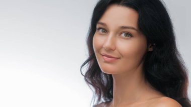 Siyah saçlı, kusursuz tenli, gülümseyen genç bir kadının yakın plan portresi, yumuşak beyaz bir arka planda. Güzellik, cilt bakımı, sağlık ve doğal yaşam tarzı kavramları için mükemmel..