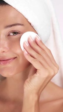Cildi ışıl ışıl parlayan genç bir kadın yüzünü pamukla silerken gülümsüyor. Kişisel bakım, cilt bakımı, temiz güzellik, minimalist sağlık ve yüz temizleme kavramları için idealdir..