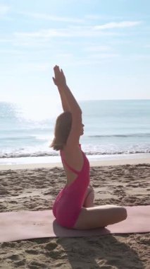 Güzel, ince bir kadın plajda tek parça pembe bir elbiseyle yoga yapıyor. Refah, vücut bakımı, güzellik, lüks geri çekilme veya fitness yaşam tarzı markalaşması için yavaş çekim konsepti.
