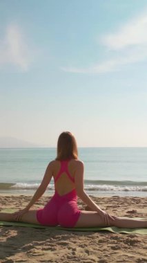 Pembe elbiseli atletik genç kadın, dağları ve okyanusu olan güneşli bir sahilde yoga yapıyor. Sağlık, esneklik, geri çekilme veya motivasyon içeriği için ideal.