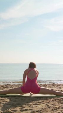 Pembe mayo giymiş zinde bir kadın plaj paspası üzerinde tam ortadan ikiye bölünüyor ve arka planda sakin bir deniz var. Esneklik, sağIık ve esneme için mükemmel..