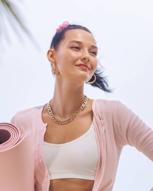 Yoga minderiyle gülümseyen genç bir kadın, günlük kıyafetlerini giymiş, kendinden emin ve açık havada rahat görünüyor. Formunu, sağlığını, yoga alışkanlıklarını veya sağlıklı yaşam tarzını geliştirmek için mükemmel bir imaj.