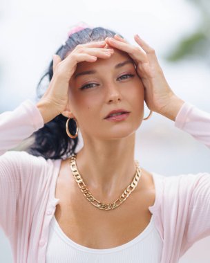 Parlayan yüzünü çerçeveleyen, altın takılar takan, güzellik, cilt bakımı ve moda reklamcılığı için ideal yumuşak pembe bir elbise giyen, açık havada poz veren beyaz bir model..