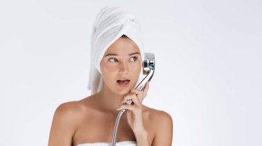 Havlu ve türbanlı beyaz kadın duş başlığını şaşırmış bir telefon gibi kulağına tutuyordu. Beyaz arkaplan ve metin için yeri olan eğlenceli güzellik ya da hijyen reklamı.