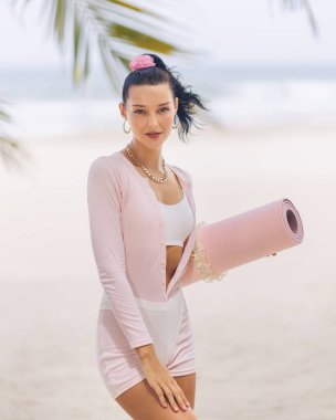 Aktif kıyafetli, elinde pembe yoga minderiyle tropik bir plajda duran şık bir kadın. Huzur ortamı ve şık görünüşü bu görüntüyü sağlık, sağlık ve yaşam tarzı terfileri için ideal kılıyor..