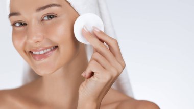 Güzel beyaz bir kadın yanağını pamukla temizlerken ışıl ışıl gülümsüyor. Havluya sarılı ve ışıl ışıl, cilt bakımı, güzellik, sağlık ve yüz ürünü reklamları için mükemmel..