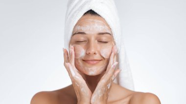 Havluya sarılı barışçıl bir kadın cilt bakımının keyfini çıkarırken yüzüne hafifçe dokunur. Işıl ışıl cildi ve gevşek ifadesi özgüveninin, güzelliğinin ve sağlığının altını çiziyor.. 