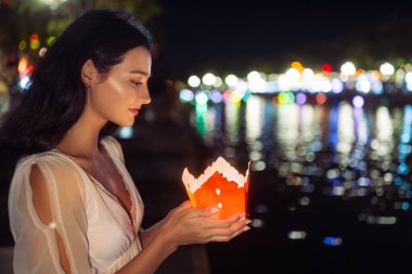 Beyaz elbiseli sakin bir kadın Hoi An 'da bir dilek tutma geleneği sırasında nehir kenarında parlayan bir kağıt fener tutuyor. Kültür, yaşam tarzı ve seyahat stoku kullanımı için harika..