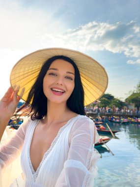 Geleneksel konik şapkalı gülümseyen kadın Vietnam 'daki Hoi An nehrinin kıyısında selfie çekiyor. Yüksek kaliteli yaşam tarzı seyahat fotoğrafı, turizm, tanıtım içeriği veya kültürel yayın kullanımı için ideal.