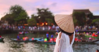 Beyaz elbiseli ve konik şapkalı beyaz bir kadının canlı Hoi An nehrine ağır çekimde bakışları. Seyahat, romantizm, özgürlük ve yaşam tarzı temaları için kavramsal görüntüler.
