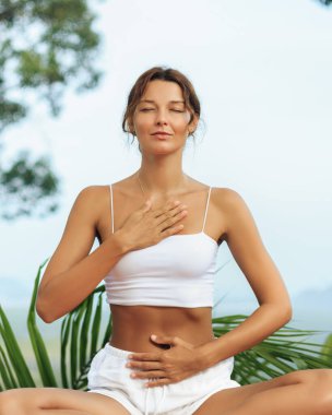 Genç bir kadın gözleri kapalı ve elleri göğsünde ve karnında dinleniyor. Tropikal doğada nefes alma farkındalığı uyguluyor. Sağlık, farkındalık ve ruhani yaşam tarzı görselleri için idealdir..
