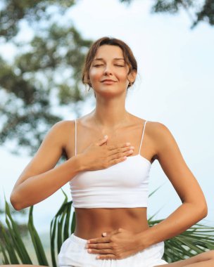 Genç kadın tropikal doğa üzerine meditasyon yapıyor elleri göğsünde ve karnında dinleniyor, gözleri kapalı ve yüzü dingin. Nefes işi, duygusal iyileşme ve sağlık temalı içerik için mükemmel..