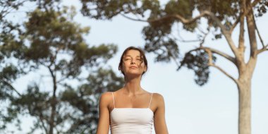 Huzurlu bir gülümsemeyle meditasyon yapan genç bir kadının ön görüntüsü ve güneşli bir orman ortamında kapalı gözler. Sağlık, farkındalık, doğa yaşam tarzı ve ruhsal geri çekilme için mükemmel..