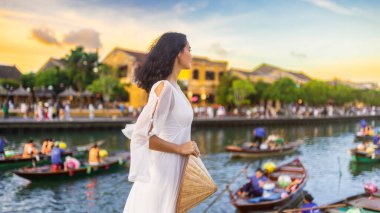 Uçan beyaz elbiseli güzel bir Asyalı kadın elinde konik bir şapkayla duruyor, Hoi An, Vietnam 'da fener dolu bir nehire bakıyor - seyahat, moda ve kültür görselleri kavramı.