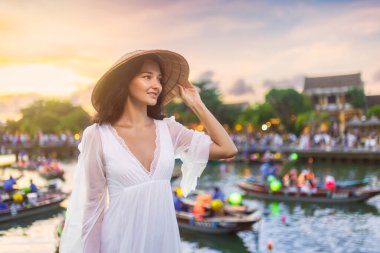 Güneş batarken Hoi An, Vietnam 'da fener ışığında gülümseyen akıcı beyaz elbiseli ve konik şapkalı hoş bir kadın. Yaşam tarzı blogları, seyahat promosyonları ve kültür kampanyaları için ideal.