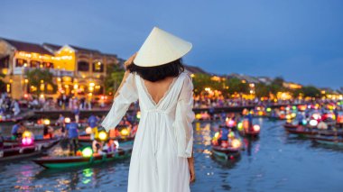 Beyaz elbiseli zarif bir kadın ve Hoi An, Vietnam 'da nehir kıyısında fenerle aydınlatılmış teknelere tepeden bakan geleneksel konik şapka. Seyahat, moda ve kültürel görseller için ideal..