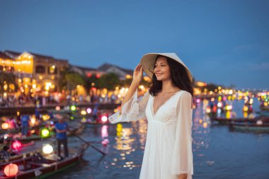 Genç Asyalı kadın Hoi An, Vietnam 'da fener ışığında eğleniyor. Beyaz bir elbise ve geleneksel bir şapka giymiş. Seyahat reklamları, kültürel içerik ve yaşam tarzı tanıtımları için mükemmel..