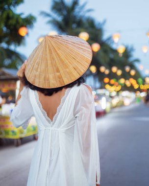 Beyaz elbiseli ve konik şapkalı şık bir kadının arka planı Hoi An 'ın fener ışığında gezen pazar caddesinde yürümesi. Seyahat reklamları, kültürel kavramlar ve borsa görüntüleri için ideal..