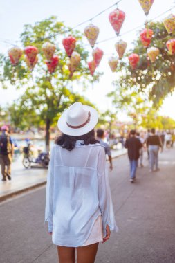 Beyaz tişörtlü ve şapkalı şık bir kadının Hoi An, Vietnam 'da renkli fenerlerin altında yürüyüşü. Rüya gibi, rahat bir seyahat anı turizm, yaşam tarzı ve blog içeriği için mükemmel.