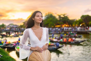 Hoi An, Vietnam 'da nehrin kenarında duran güzel genç bir kadın gün batımında konik olmayan geleneksel bir şapka tutuyor. Sahnede renkli tekneler, yumuşak altın ışık ve huzurlu bir akşam var.
