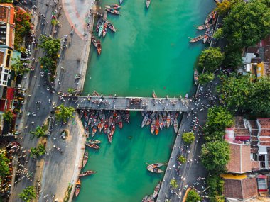Hoi An, Vietnam 'daki zümrüt yeşili nehrin üzerindeki yaya köprüsünün yanında toplanmış geleneksel teknelerin çarpıcı hava fotoğrafı. Seyahat, kültür ve editoryal görseller için mükemmel..