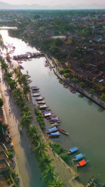 İnsansız hava aracı görüntüleri, Hoi An, Vietnam 'daki Thu Bon nehri boyunca demirlemiş palmiye çizgili nehir kenarının ve teknelerin dikey görüntülerini gösteriyor. Kültür, turizm ve yaşam tarzı görselleri için ideal.