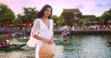 Beyaz elbiseli zarif kadın konik şapkasını tutuyor ve Hoi An 'da pembe bir gökyüzünün altında nehri seyrediyor. Romantik yavaş çekim anı seyahat, moda ve lüks yaşam tarzı medyası için mükemmel.